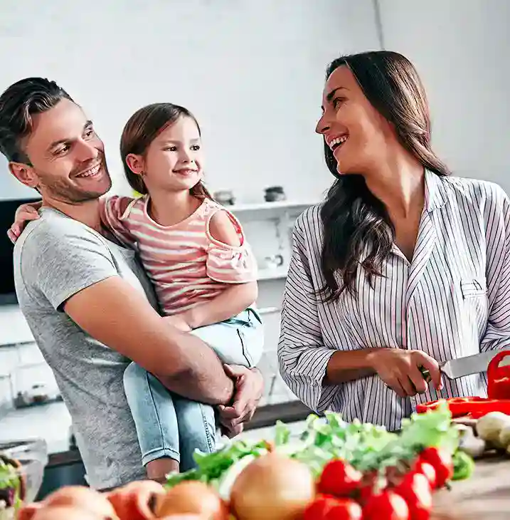 A man holds a smiling young girl while a woman chops vegetables at a kitchen counter. All three are smiling and appear happy, surrounded by fresh produce.