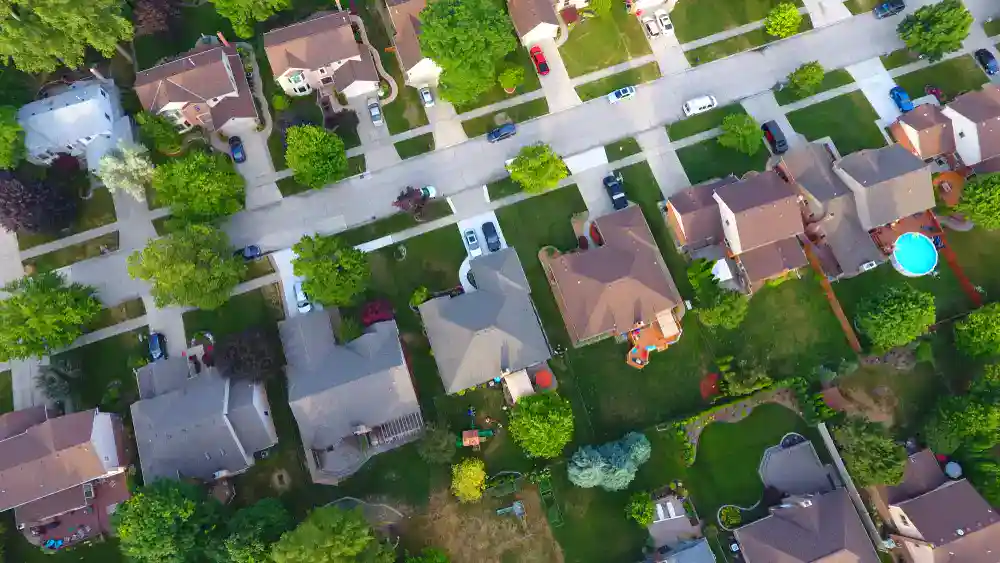 Aerial view of a suburban neighborhood showing houses with driveways, green lawns, trees, and backyards. One backyard has a round blue swimming pool visible in the lower right corner.