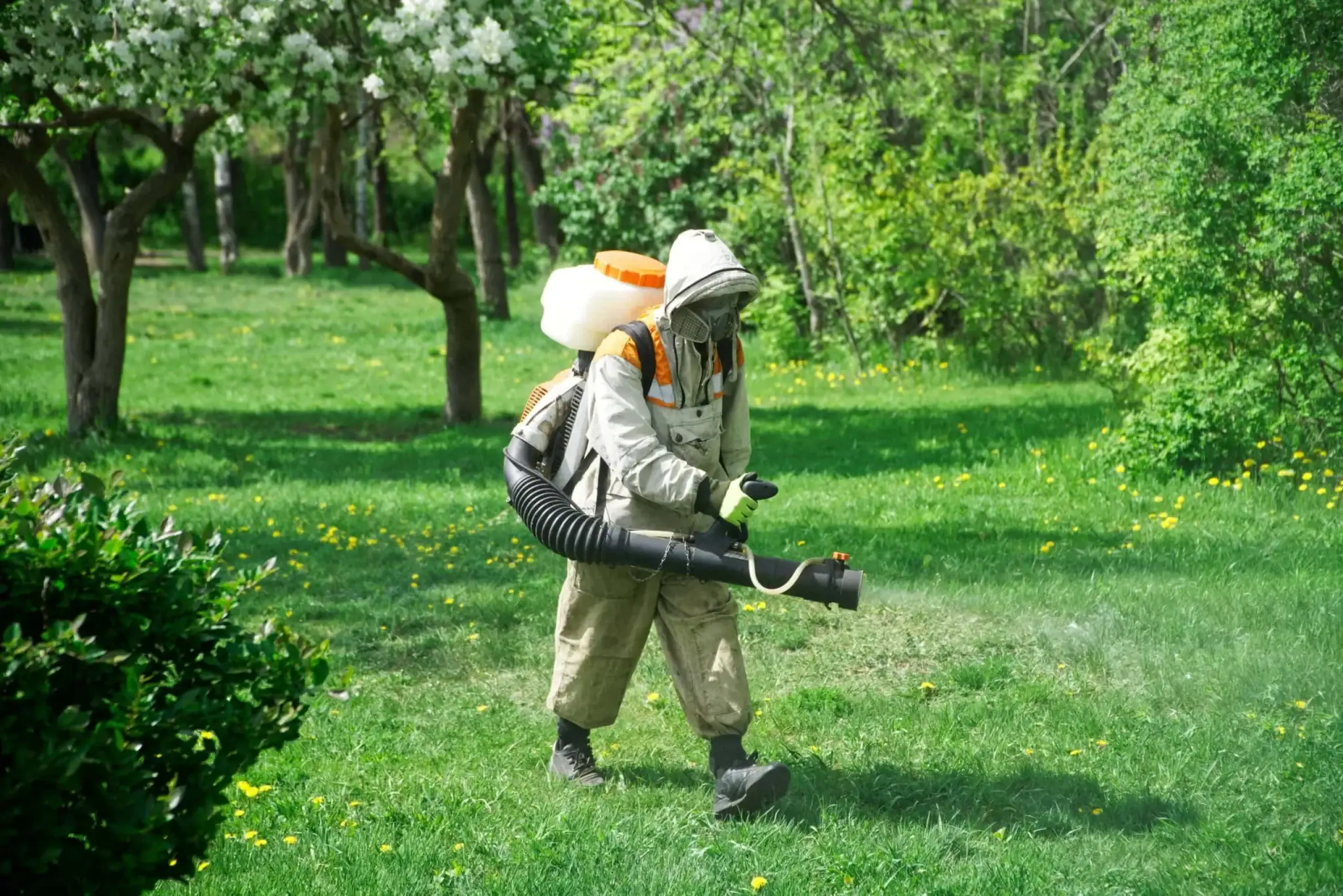 A person wearing protective gear sprays chemicals on a grassy lawn with trees and bushes in the background, likely performing pest control in Genesee County, MI.